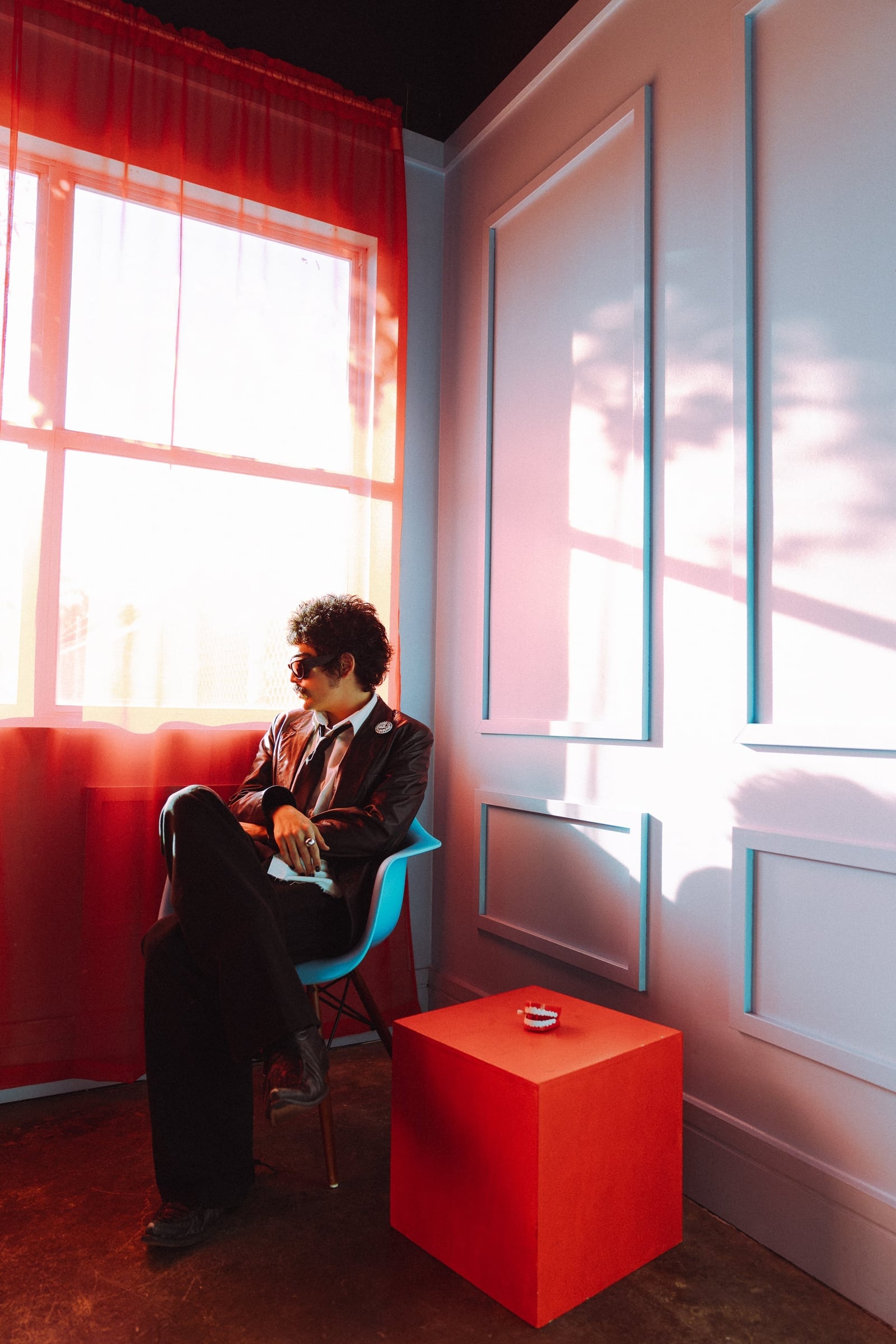 Almost Heaven — editorial portrait of the artist seated by a red-curtained window in a leather jacket and tie, dramatic red light across a paneled wall — editorial music photography by Chauncey Scott, Austin TX