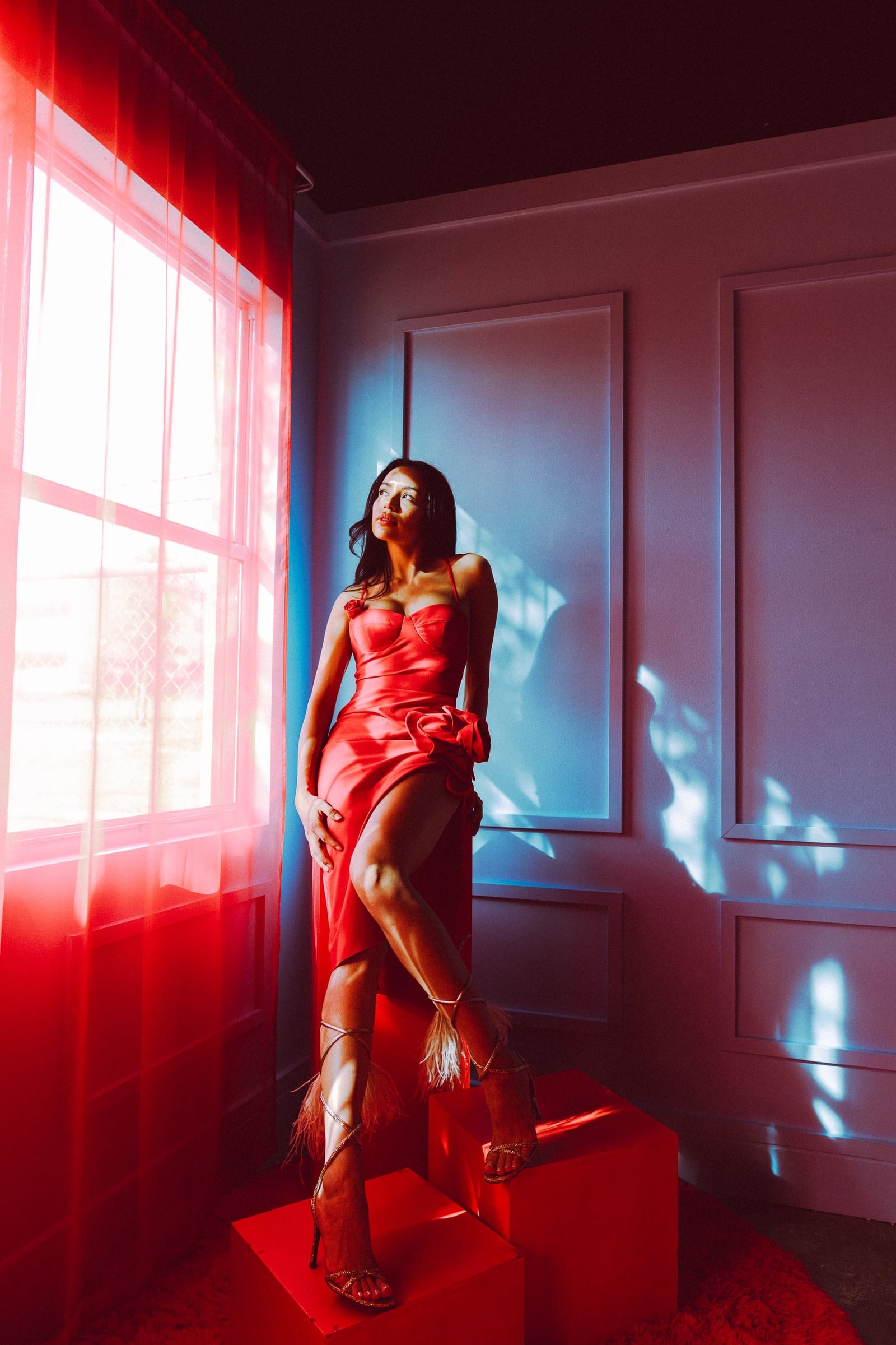 Aly Zuniga in a red satin rosette gown posed on red blocks beside a sheer curtain, color fashion editorial with red gel light by Chauncey Scott, Austin TX