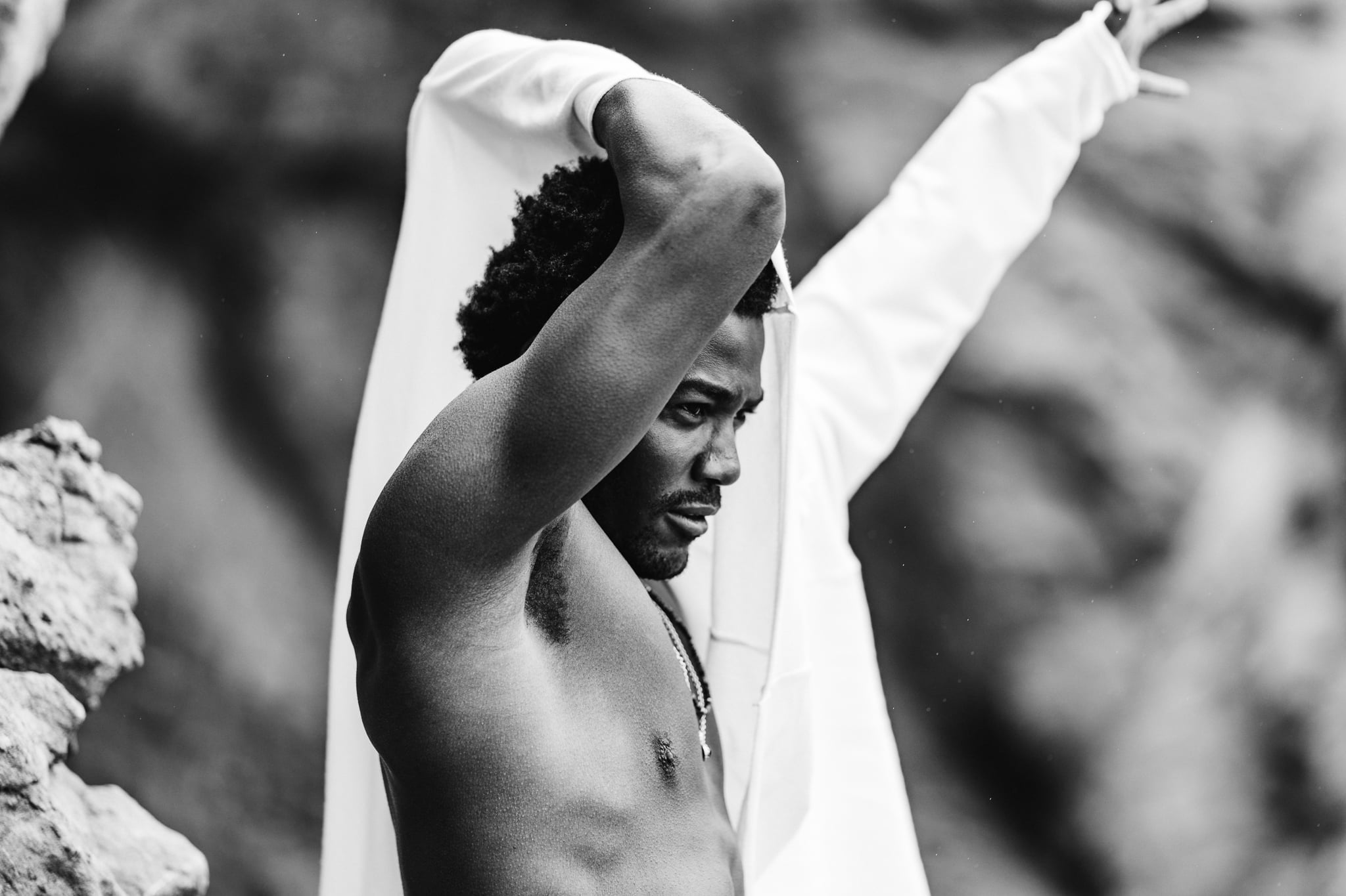 Copala model raising his arms in a white shirt against the Malibu rocks, black-and-white menswear lifestyle editorial by Chauncey Scott, Leo Carrillo State Beach