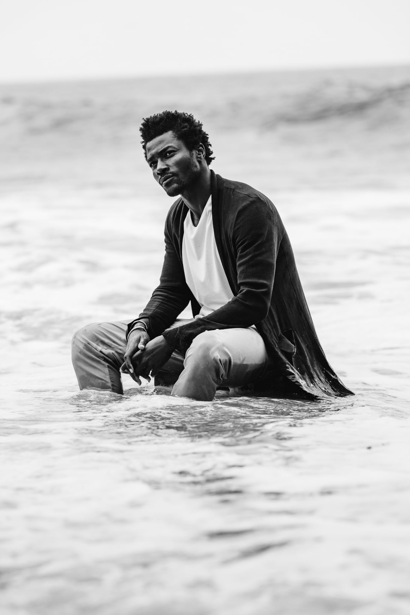Copala model crouched in the breaking surf in a charcoal blazer and white tee, black-and-white menswear lifestyle editorial by Chauncey Scott, Malibu CA