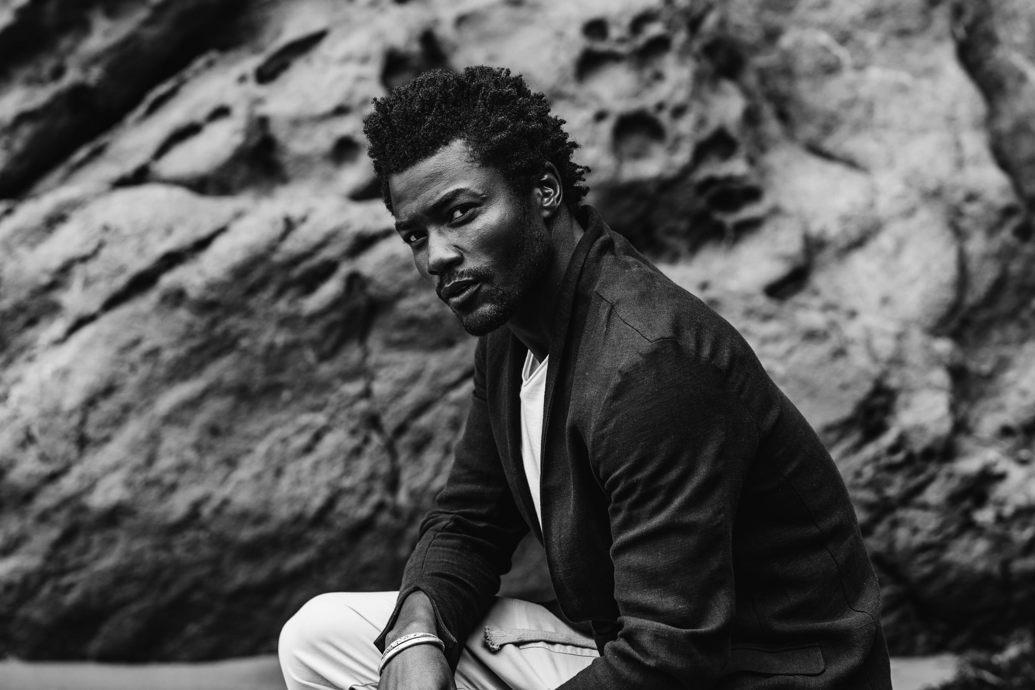 Copala model seated on a boulder in a charcoal blazer and white tee, black-and-white menswear editorial by Chauncey Scott, Leo Carrillo State Beach, Malibu
