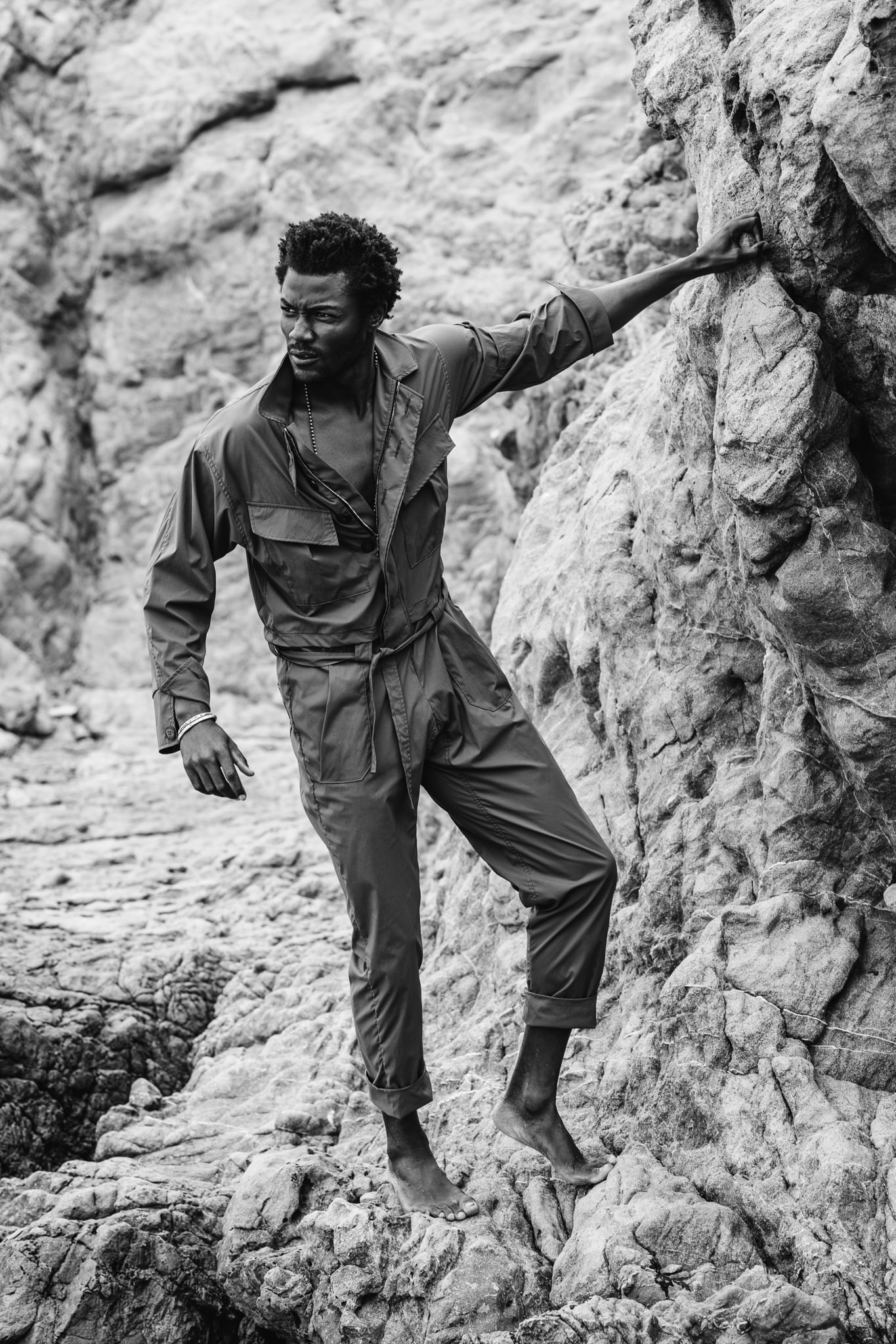 Copala model climbing barefoot up coastal rocks in an olive utility coverall, black-and-white menswear lifestyle editorial by Chauncey Scott, Malibu CA