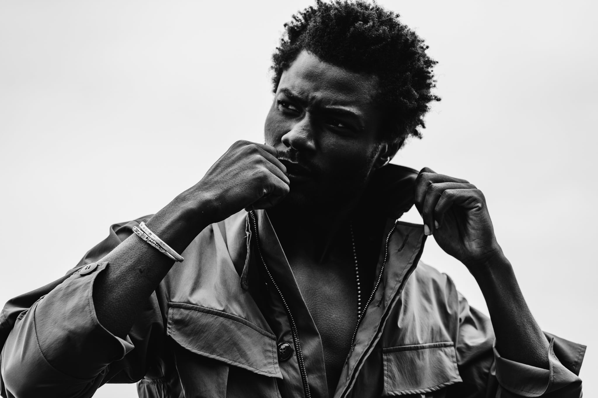 Copala model gripping the collar of an olive utility jacket against a bright sky, black-and-white menswear portrait by Chauncey Scott, Malibu CA