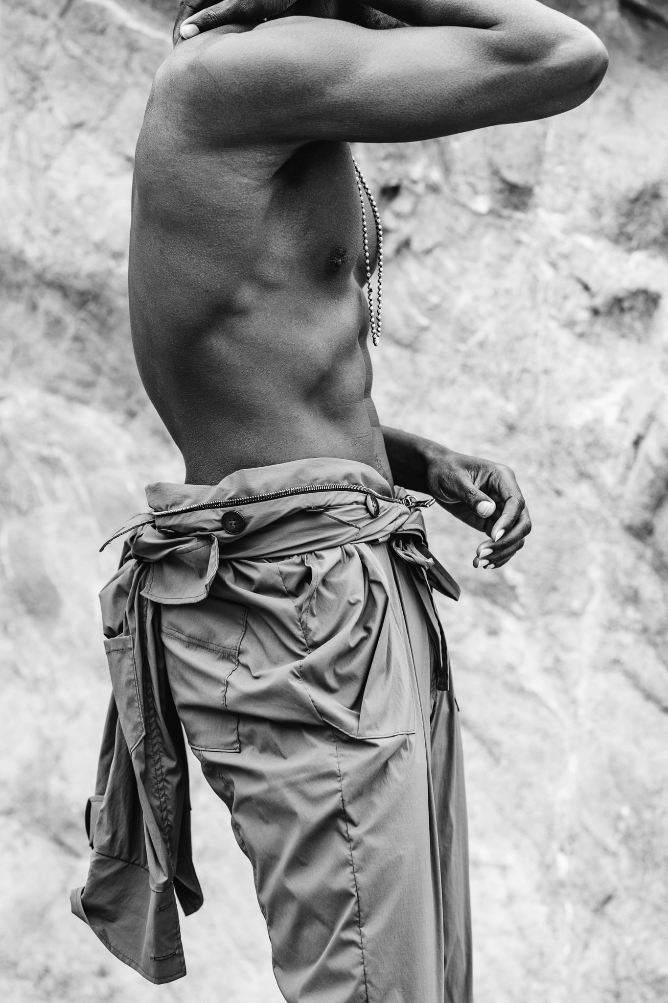 Copala model shirtless in profile in olive cargo pants with a jacket tied at the waist, black-and-white menswear editorial by Chauncey Scott, Malibu CA