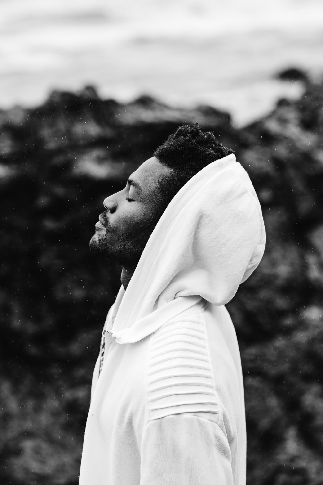 Copala model in profile with eyes closed in a white hooded jacket amid sea spray, black-and-white menswear editorial by Chauncey Scott, Malibu CA