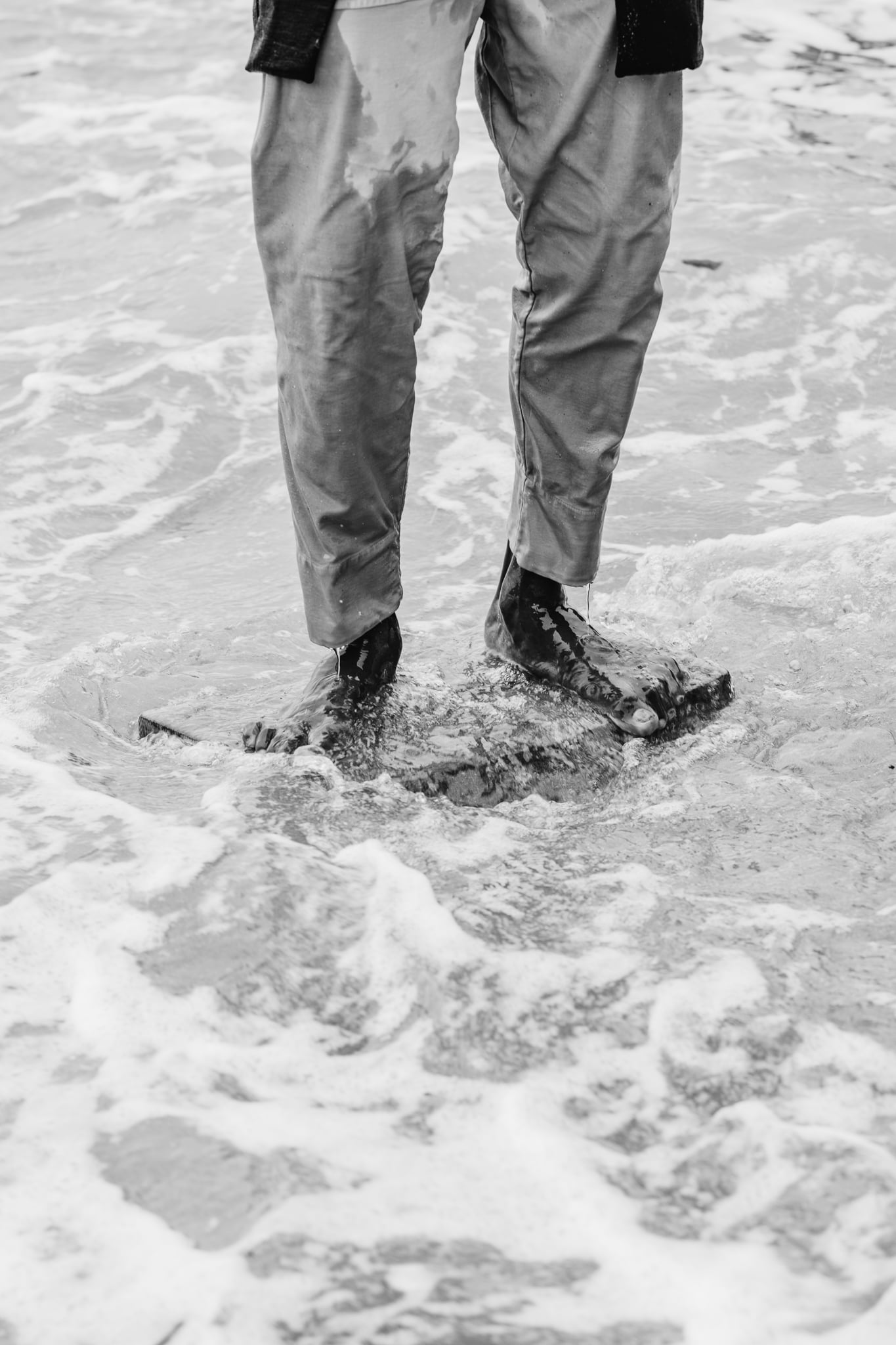 Copala model standing barefoot in foaming surf with trousers soaked, black-and-white menswear editorial by Chauncey Scott, Leo Carrillo State Beach