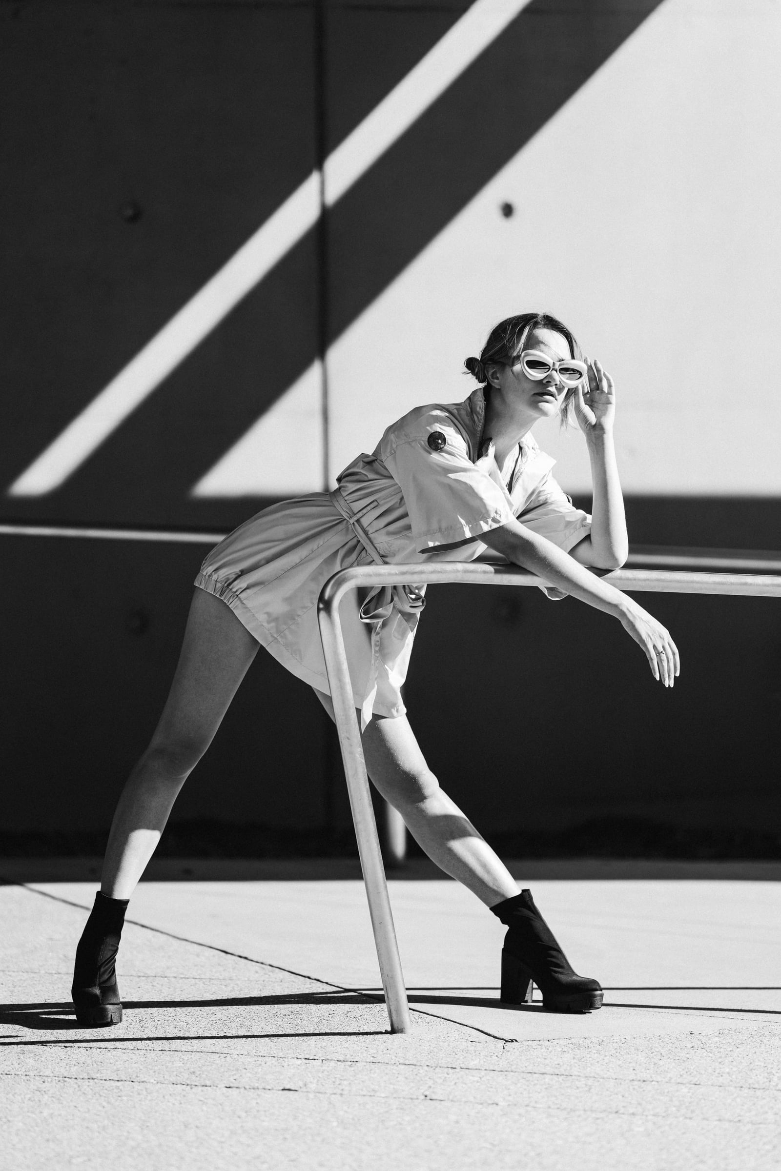 Devin Thomas in a belted utility shirtdress and white oval sunglasses, leaning on a steel handrail against the sunlit concrete facade of the Perot Museum, Dallas, Texas — fashion editorial by Chauncey Scott