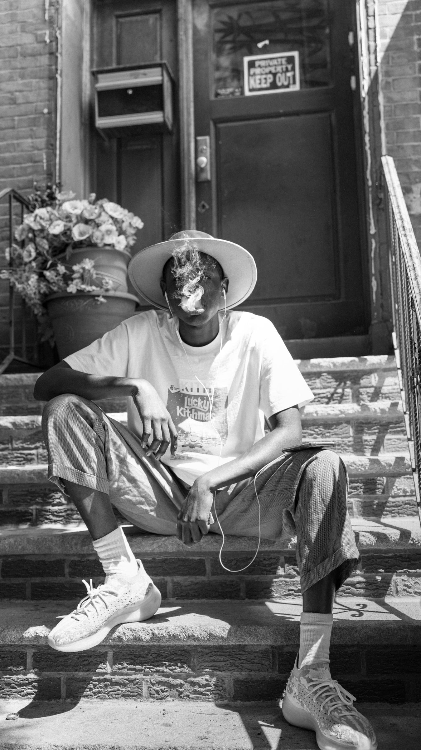 Black-and-white editorial portrait — Joe West seated on a Brooklyn brownstone stoop in a wide-brim hat and graphic tee, exhaling smoke beside a pot of flowers — portrait photography by Chauncey Scott