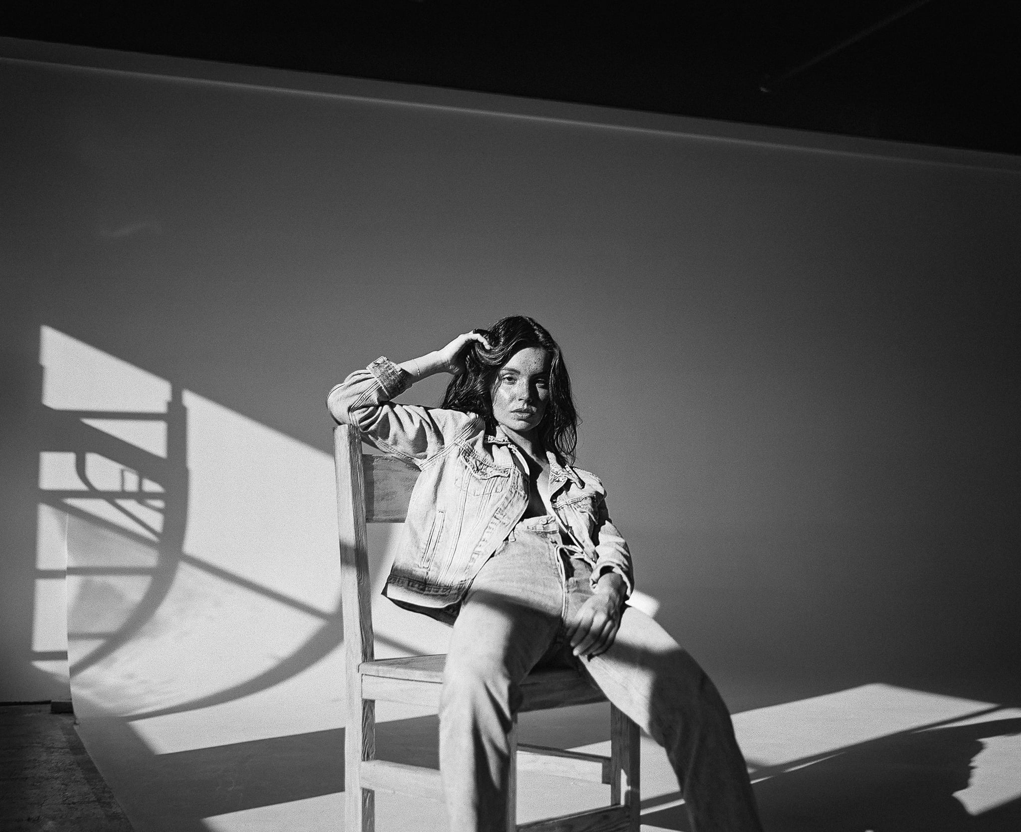 Lillian Connell seated on a wooden chair in a Levi's denim jacket and jeans as a window-frame shadow falls across the wall, black-and-white editorial by Chauncey Scott, Austin TX