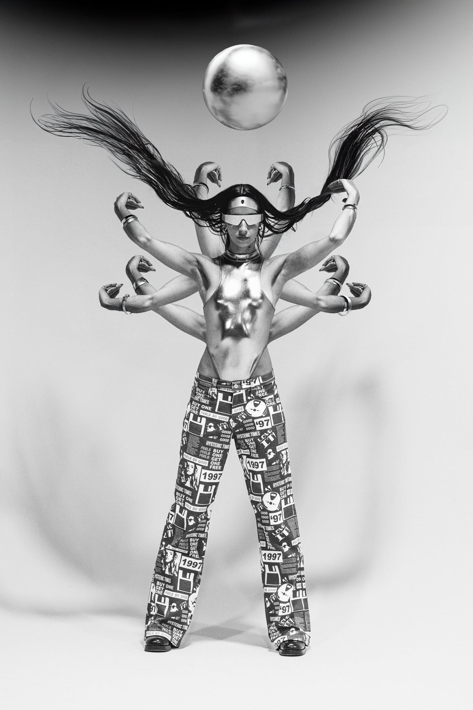 Black-and-white studio fashion editorial — a model in a chrome bodysuit and visor with multiple arms fanned out and hair streaming upward beneath a floating silver orb, in graphic-print flared pants — fashion photography by Chauncey Scott