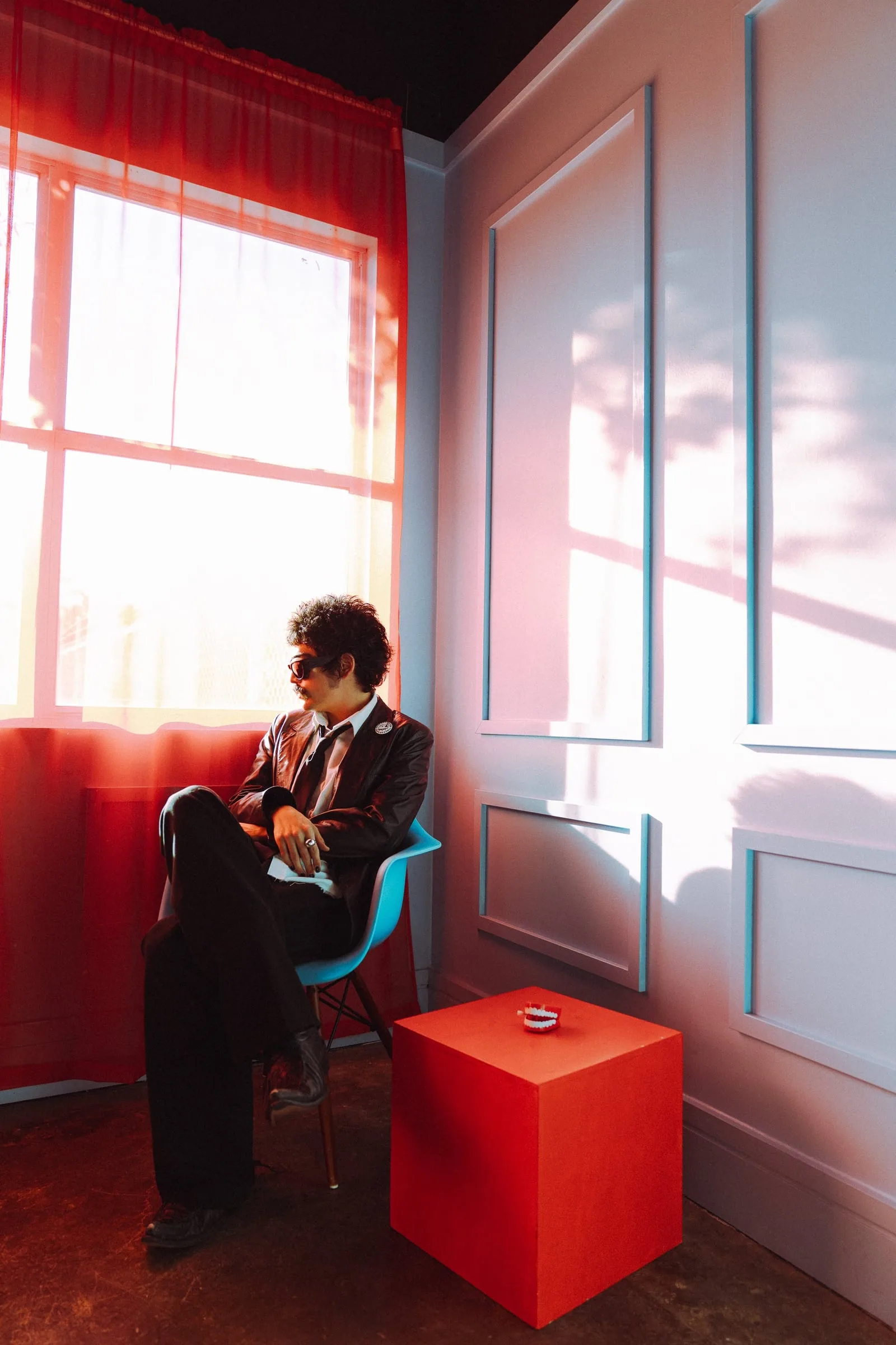 Almost Heaven — editorial portrait of the artist seated by a red-curtained window in a leather jacket and tie, dramatic red light across a paneled wall — editorial music photography by Chauncey Scott, Austin TX